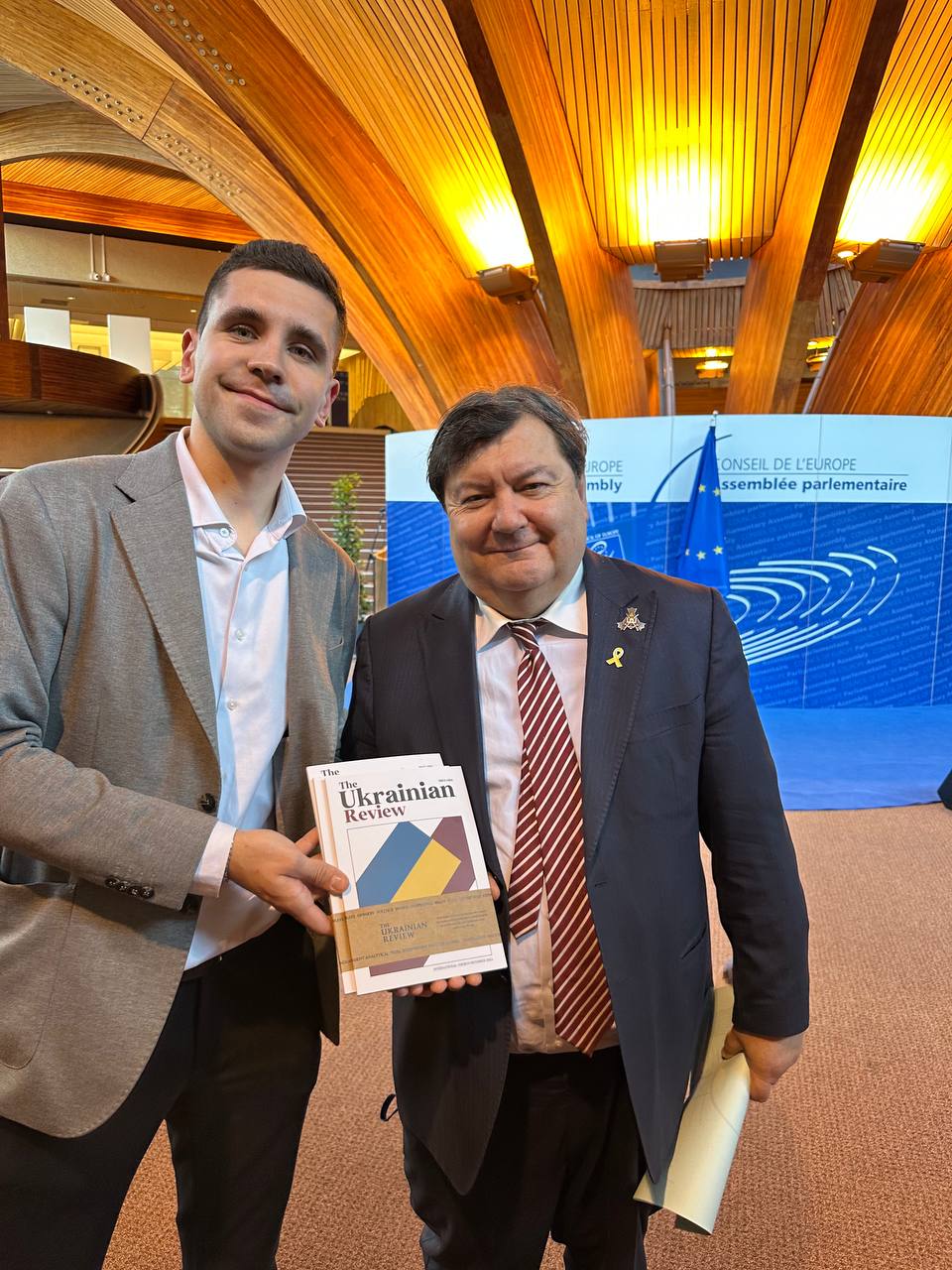 Two PACE delegates holding The Ukrainian Review in front of the Council of Europe backdrop, April 2025.