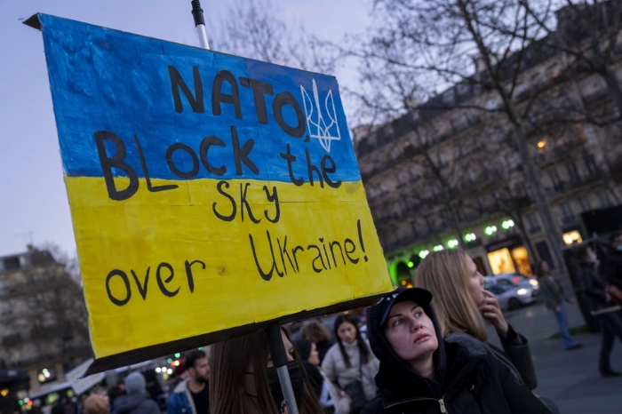 A protest sign painted in the colors of the Ukrainian flag reads “NATO block the sky over Ukraine!” during a public demonstration. People are gathered in the background on a city street, showing support for Ukraine and calling for international action amid the war.