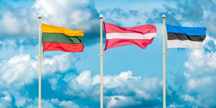 Three national flags of the Baltic States — Lithuania, Latvia, and Estonia — wave on flagpoles against a bright blue sky with clouds.