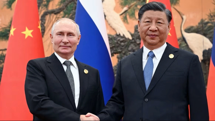 Russian President Vladimir Putin and Chinese leader Xi Jinping are pictured shaking hands during a bilateral meeting in Beijing in October 2023. Both leaders appear in dark business suits and ties against the backdrop of Chinese and Russian national flags, underscoring the symbolic display of their partnership.