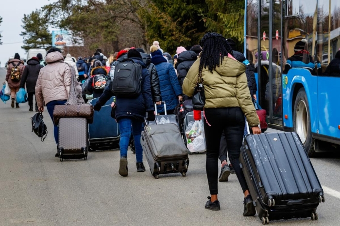 The image shows a group of people walking along a road with suitcases and bags, many wearing warm clothing, as they move toward or away from buses. It conveys the scene of Ukrainian refugees