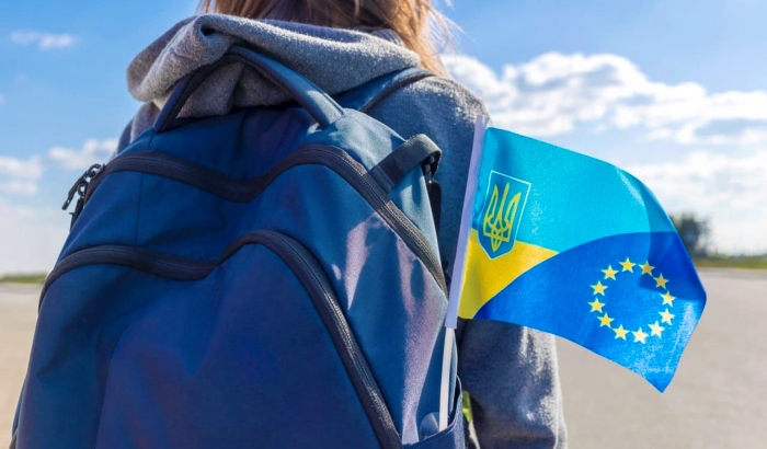 The photo shows a person wearing a blue backpack with a small flag tucked into it. The flag combines Ukraine’s national colors (blue and yellow with the trident coat of arms) and the European Union’s circle of stars, symbolizing Ukraine’s aspiration for closer ties with Europe. The image conveys movement, youth, and mobility, suggesting themes of travel, education abroad, return migration, or integration with the EU.