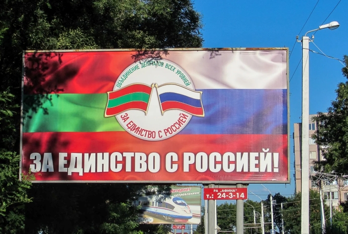 The image shows a large outdoor billboard in a city promoting pro-Russian unity in Transnistria. The billboard features the flags of Transnistria (green-red) and Russia (white-blue-red) crossed in the center with a circular emblem reading “For Unity with Russia” in Russian. Below, in bold white Cyrillic letters on a red background, it says “За единство с Россией!” (“For unity with Russia!”). Trees, electrical poles, and other street signs are visible around the sign.