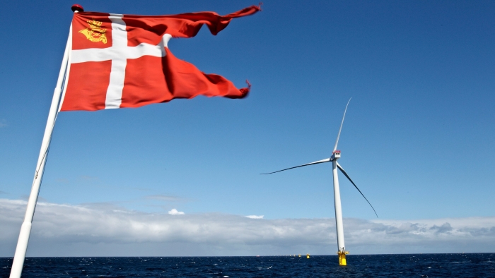 The image shows a Danish flag with a coat of arms fluttering in the foreground against a bright blue sky. In the background, there’s a large offshore wind turbine standing in the sea, symbolising Denmark’s strong focus on renewable energy.