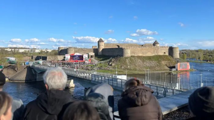 The photo shows a large group of people standing on the Estonian side of the Narva River, looking across the water toward the Russian city of Ivangorod.