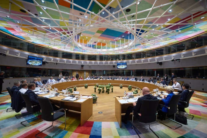 The image shows a meeting of European Union leaders at the European Council headquarters in Brussels. Delegates are seated around a large circular table beneath a colorful, geometric glass ceiling — a hallmark of the Europa Building