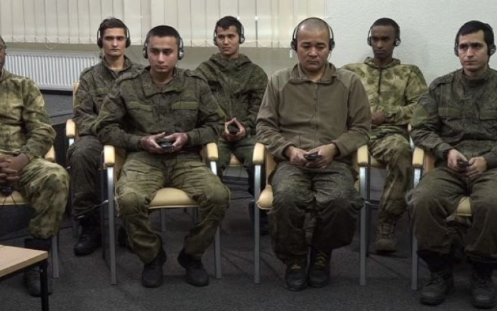 Group of Russian war prisoners from different nations are shown seated in Russian military uniforms on chairs with headphones