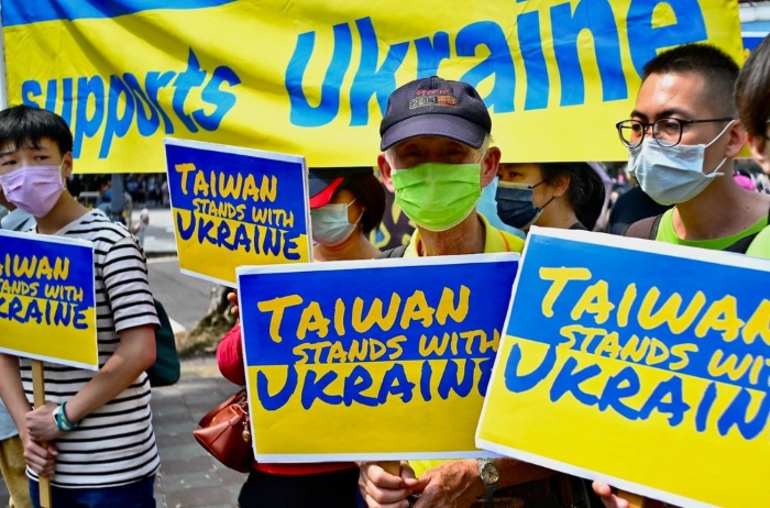 The image shows a group of people in Taiwan holding blue and yellow signs that read “Taiwan stands with Ukraine.” They are wearing face masks and standing in front of a large banner that says “supports Ukraine.” The scene depicts a solidarity rally or protest in support of Ukraine amid Russia’s invasion, symbolizing shared democratic values and opposition to aggression.