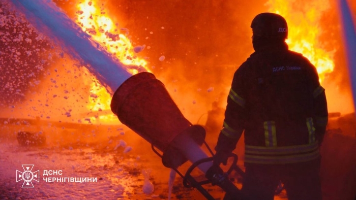 Ukrainian firefighter from the State Emergency Service battles a massive blaze with foam and water during a night fire in Chernihiv region, flames and smoke rising in the background.