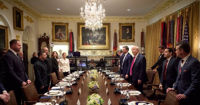 The image shows a formal meeting between Ukrainian and American delegations in an elegant, chandelier-lit room at the White House. President Volodymyr Zelenskyy and U.S. President Donald Trump stand facing each other across a long conference table set for a diplomatic dinner.
