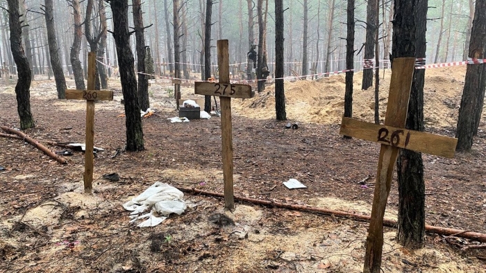 The image shows a mass burial site in a forested area near Izium, Ukraine, where wooden crosses marked with numbers stand over shallow graves. The site, discovered after Russian occupation, became evidence of war crimes and civilian killings during the invasion. The photo captures a grim scene of mist, trees, and forensic tape — symbolizing the tragedy and scale of Russian atrocities in occupied territories.