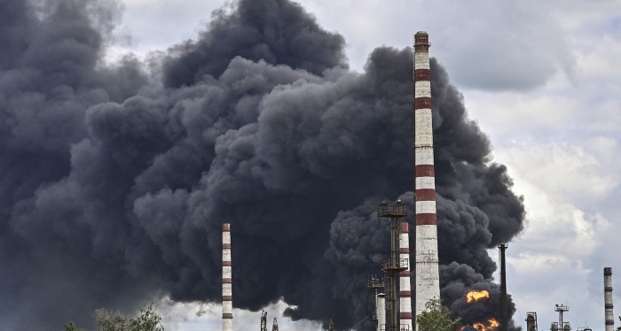 Massive black smoke rises from an industrial facility with tall chimneys, indicating a large fire or explosion at an oil refinery or chemical plant. The scene shows intense flames and pollution against a cloudy sky.