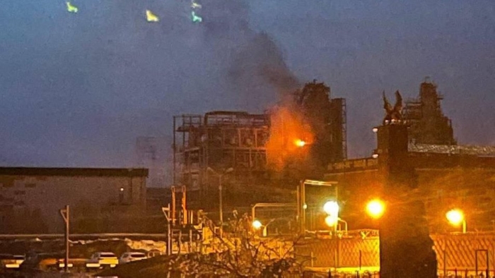 Nighttime explosion at an industrial facility in Russia with visible fire and smoke rising from the building. Emergency lights illuminate the area as flames spread across the structure. The image captures the intensity of the blaze and its impact on the surrounding site.
