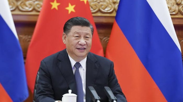 Chinese leader sits at a conference table with microphones in front of the flags of China and Russia, symbolizing close political and diplomatic ties between the two countries.