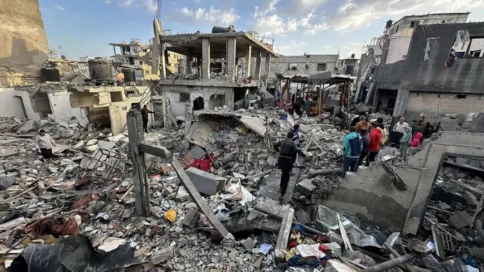 The image shows extensive destruction in a residential area of Gaza, with collapsed buildings, debris, and people searching through the rubble after heavy airstrikes.
