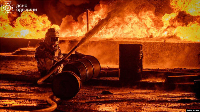 A firefighter in protective gear battles a massive blaze at an industrial site in Odesa region, Ukraine. The intense flames and smoke illuminate the night as emergency services work to contain the fire.