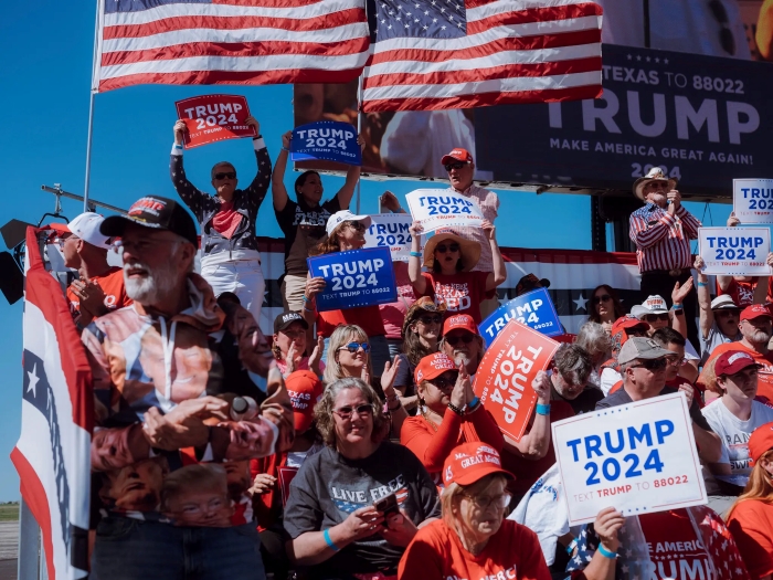 A large crowd of supporters gathers at a political rally holding “Trump 2024” signs and wearing red MAGA hats. Multiple American flags wave above the group as people cheer and display campaign posters.