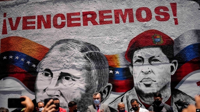 A vibrant political mural featuring Russian President Vladimir Putin and late Venezuelan President Hugo Chávez painted against the backdrop of the Venezuelan flag, with the bold slogan “¡Venceremos!” above them. People wearing masks stand in front of the artwork during a public event. The scene highlights themes of political messaging, Latin American symbolism, and street art.