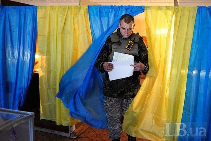 A Ukrainian servicemember in uniform exits a voting booth decorated with the national flag, holding election documents.