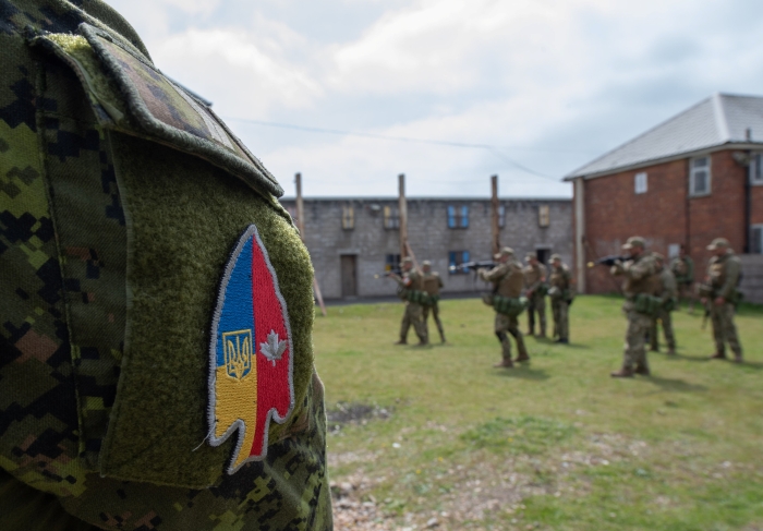 A group of soldiers in full combat gear conduct coordinated training exercises in an outdoor urban-style training area. In the foreground, a close-up uniform patch displays a combined Ukraine–Canada insignia, symbolizing joint military cooperation.