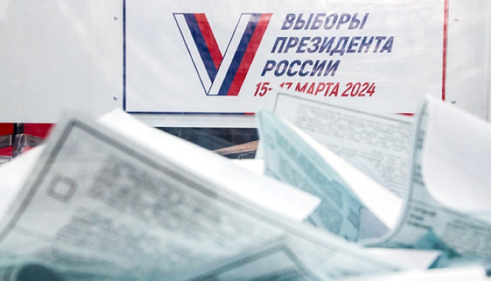 The photo shows a ballot box filled with voting papers during the 2024 Russian presidential election, with an official banner in the background displaying the voting dates, March 15–17.