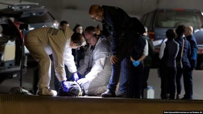The image shows a forensic response team conducting an on-site examination in what appears to be a controlled, high-security environment. Several specialists in protective suits are inspecting an item on the ground, using focused lighting and technical equipment