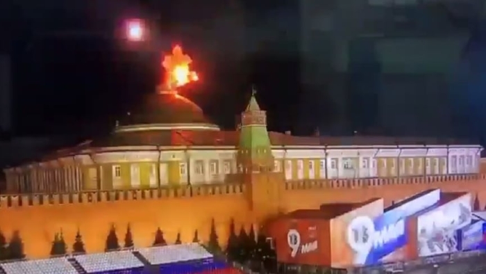 A nighttime view of a Kremlin complex building with flames visible on the rooftop, illuminating the historic structure against the dark sky.