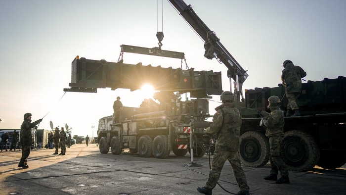 Military personnel operate heavy equipment to unload and position a missile launcher from a transport vehicle