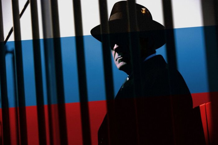 A shadowy figure in a fedora hat stands behind vertical bars, symbolizing espionage and secrecy, with the Russian flag overlaid across the entire scene. The dark silhouette and tricolor background evoke themes of Russian intelligence operations, covert agents, and geopolitical tension. This image is ideal for articles about Russian spies in Europe, Kremlin espionage networks, foreign interference, or security threats.
