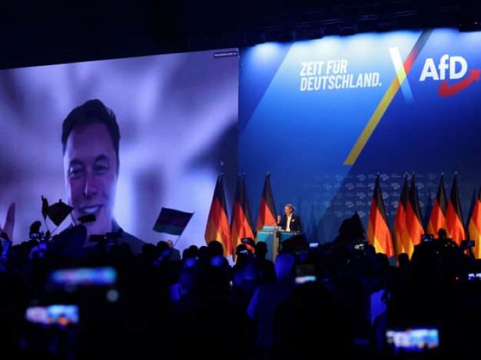 A large political rally features a massive screen with a smiling Elon Mask speaking via video call, while an AFD presenter stands at a podium on stage surrounded by multiple German flags