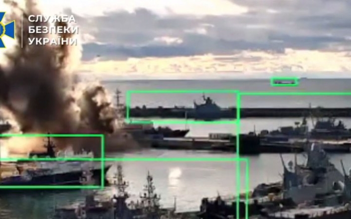 Image shows an explosion at a Russian naval harbor, with multiple military ships docked near the blast site.