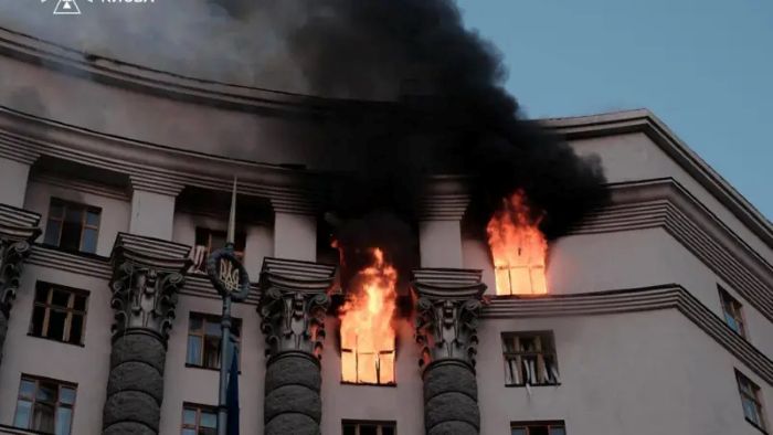 A building engulfed in flames and thick black smoke following an apparent attack, with fire visible through shattered windows. The image illustrates the aftermath of an explosion or strike on the Ukrainian government building.