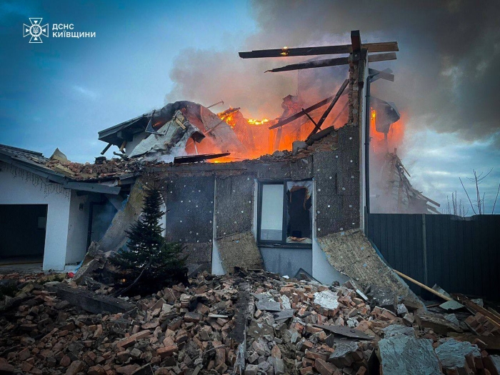 Image shows a residential building in the Kyiv region severely damaged by a russian night attack. Flames rise from the collapsed roof, with rubble, broken walls, and debris scattered around the house,