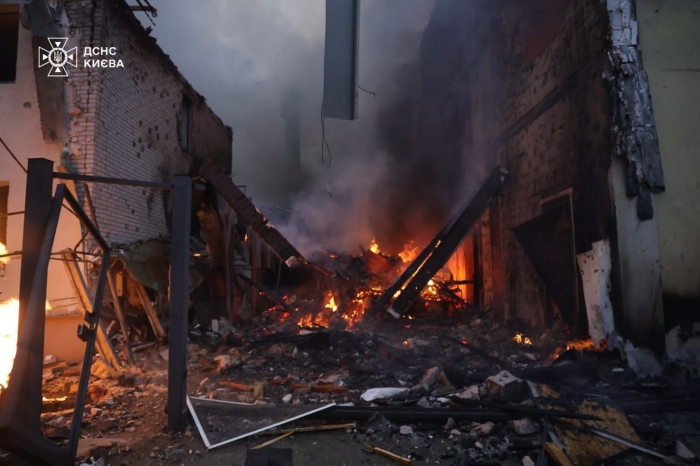 A nighttime photo showing the aftermath of a missile or drone strike in Kyiv: a heavily damaged building with collapsed walls, burning debris, and thick smoke rising from the ruins.
