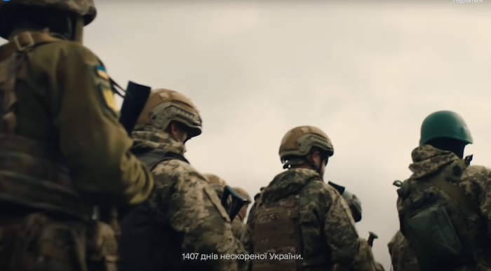 Ukrainian soldiers in combat gear stand together during a military operation, viewed from behind under an overcast sky.