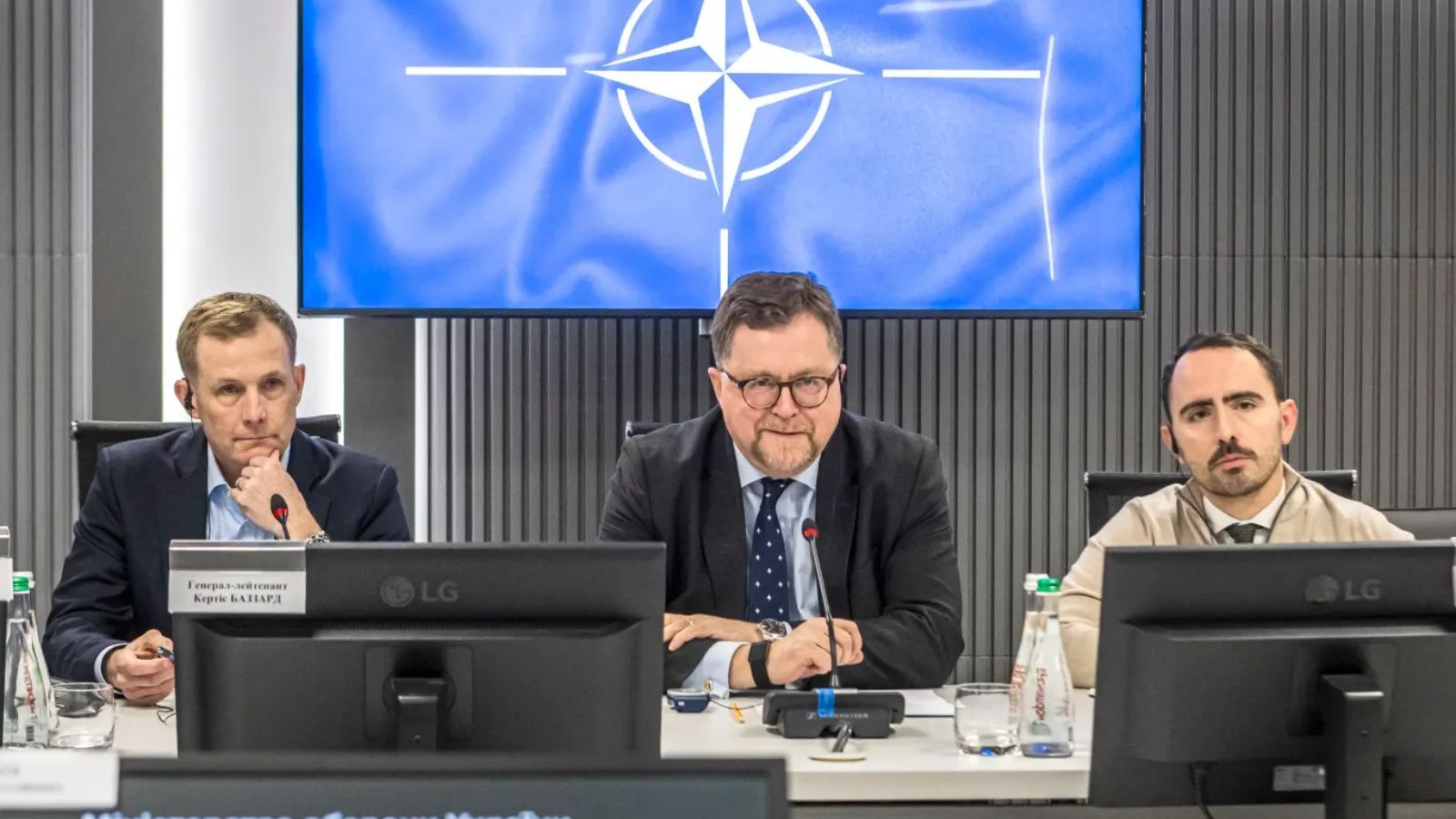 Patrick Turner and Lieutenant General Curtis Buzzard sitting at a briefing table with the NATO flag in the background.
