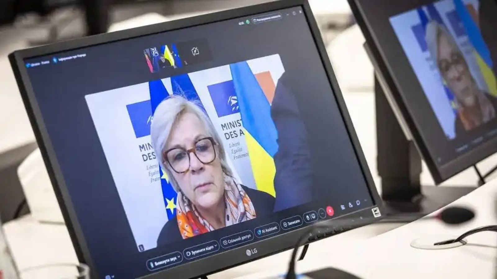 A computer monitor displaying Minister Catherine Vautrin during a video call, with Ukrainian flags visible in the background.