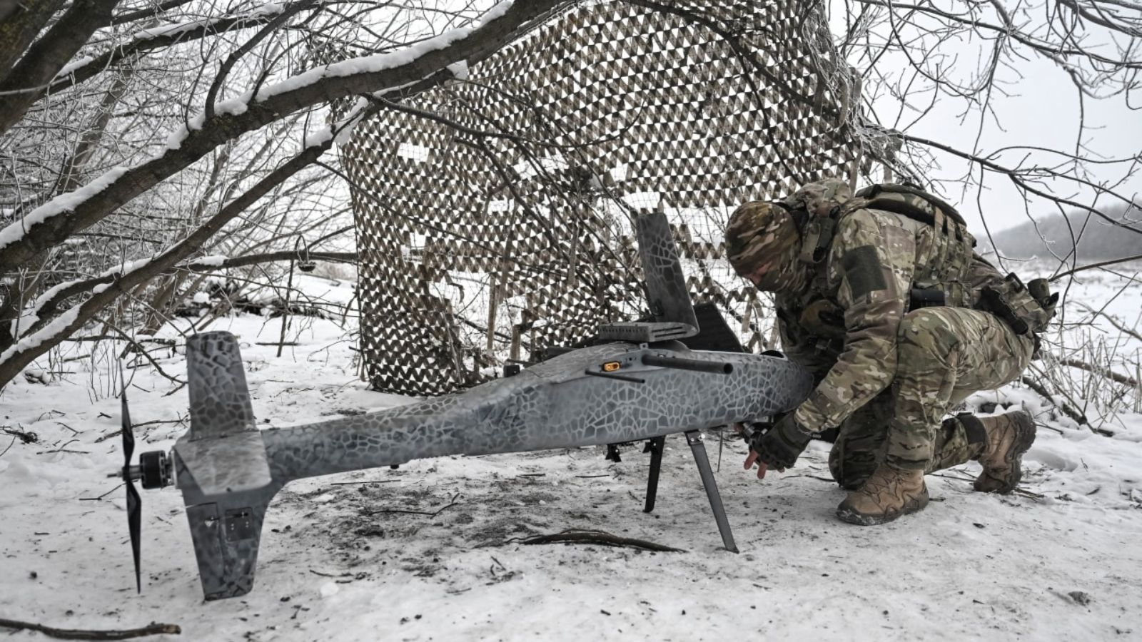 Військовий у повній амуніції налаштовує великий сірий дрон серед засніжених дерев та маскувальних сіток.