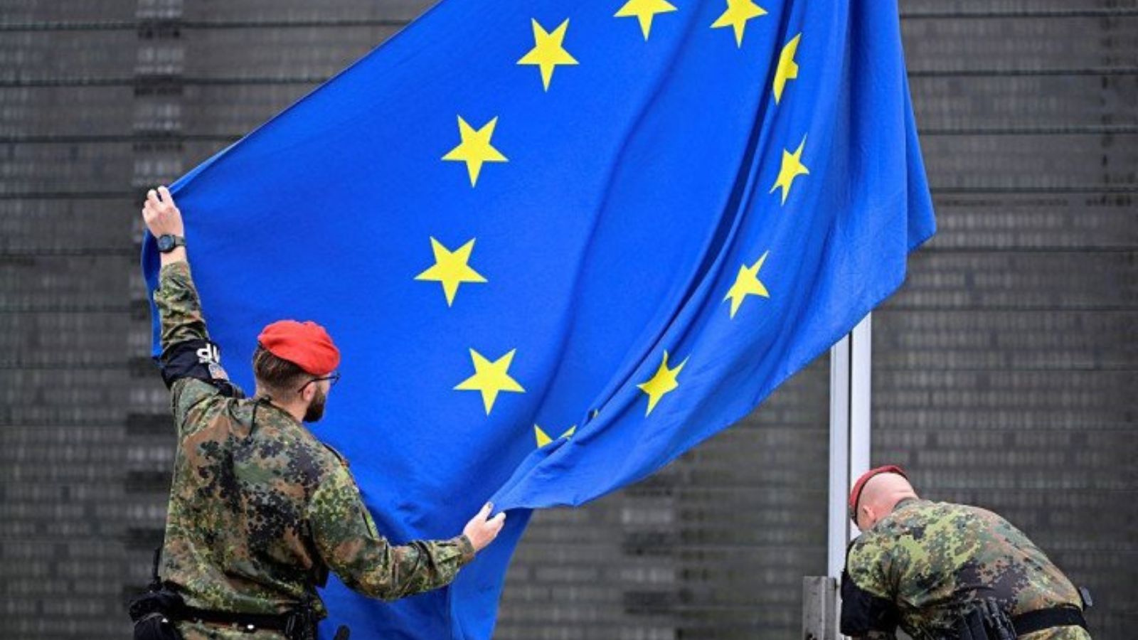 Two German soldiers in camouflage and red berets unfurling a large EU flag.