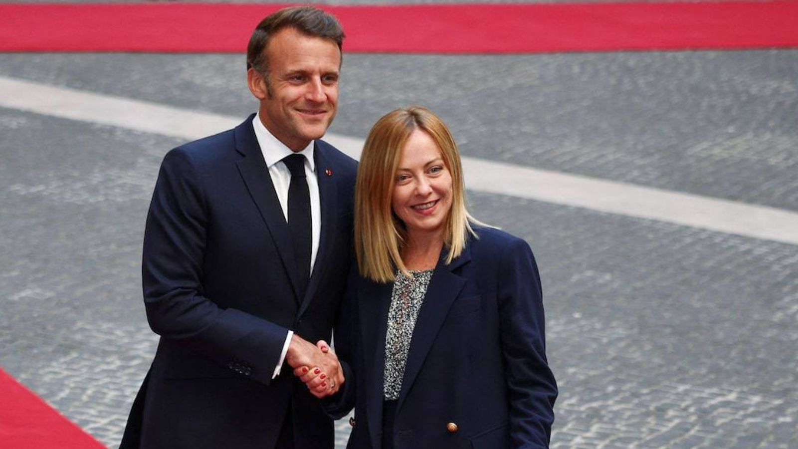 French President Emmanuel Macron and Italian Prime Minister Giorgia Meloni smiling and shaking hands while standing on a red carpet.