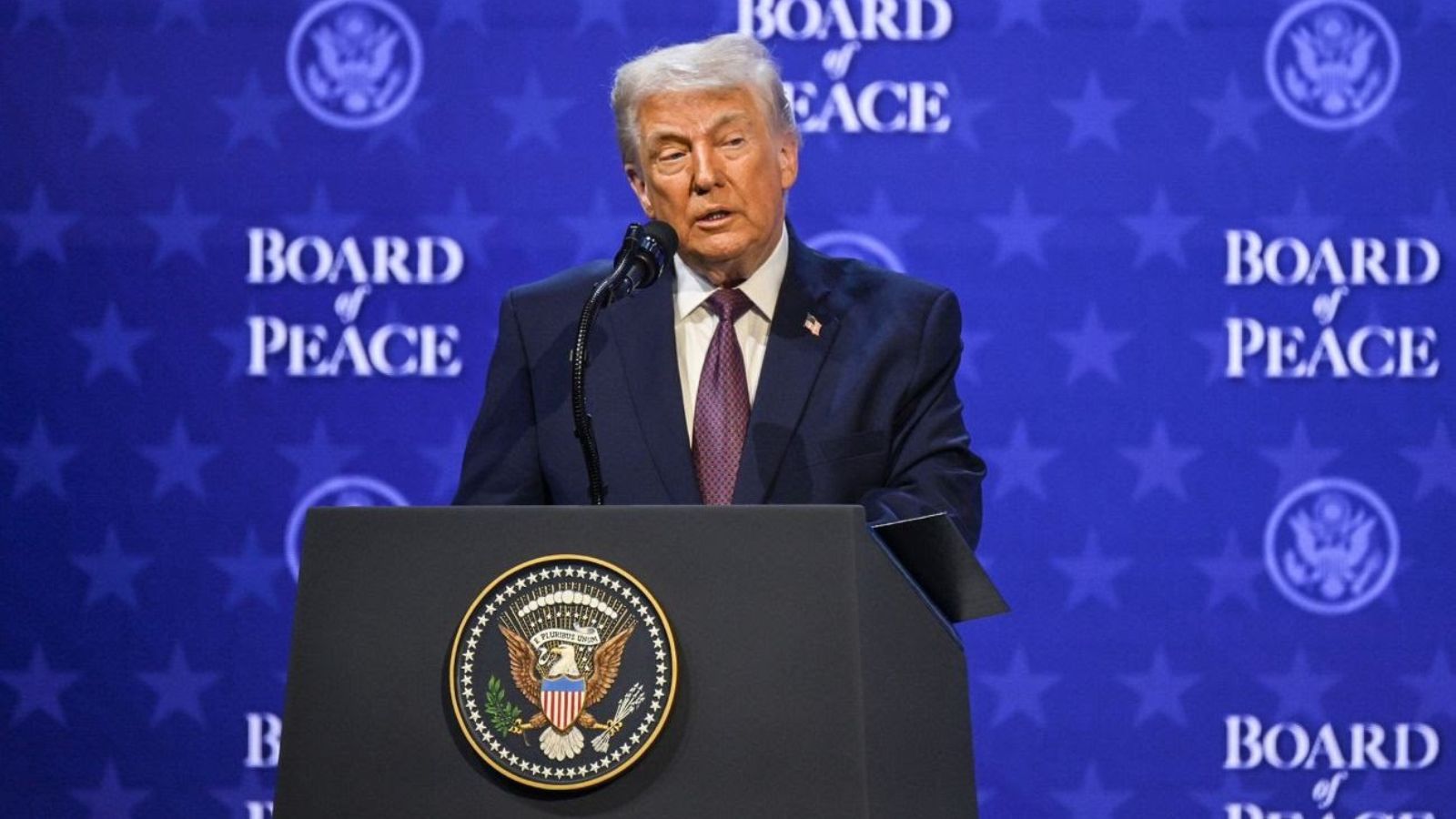Donald Trump speaking at a podium with the US Presidential Seal against a background banner that reads "Board of Peace."