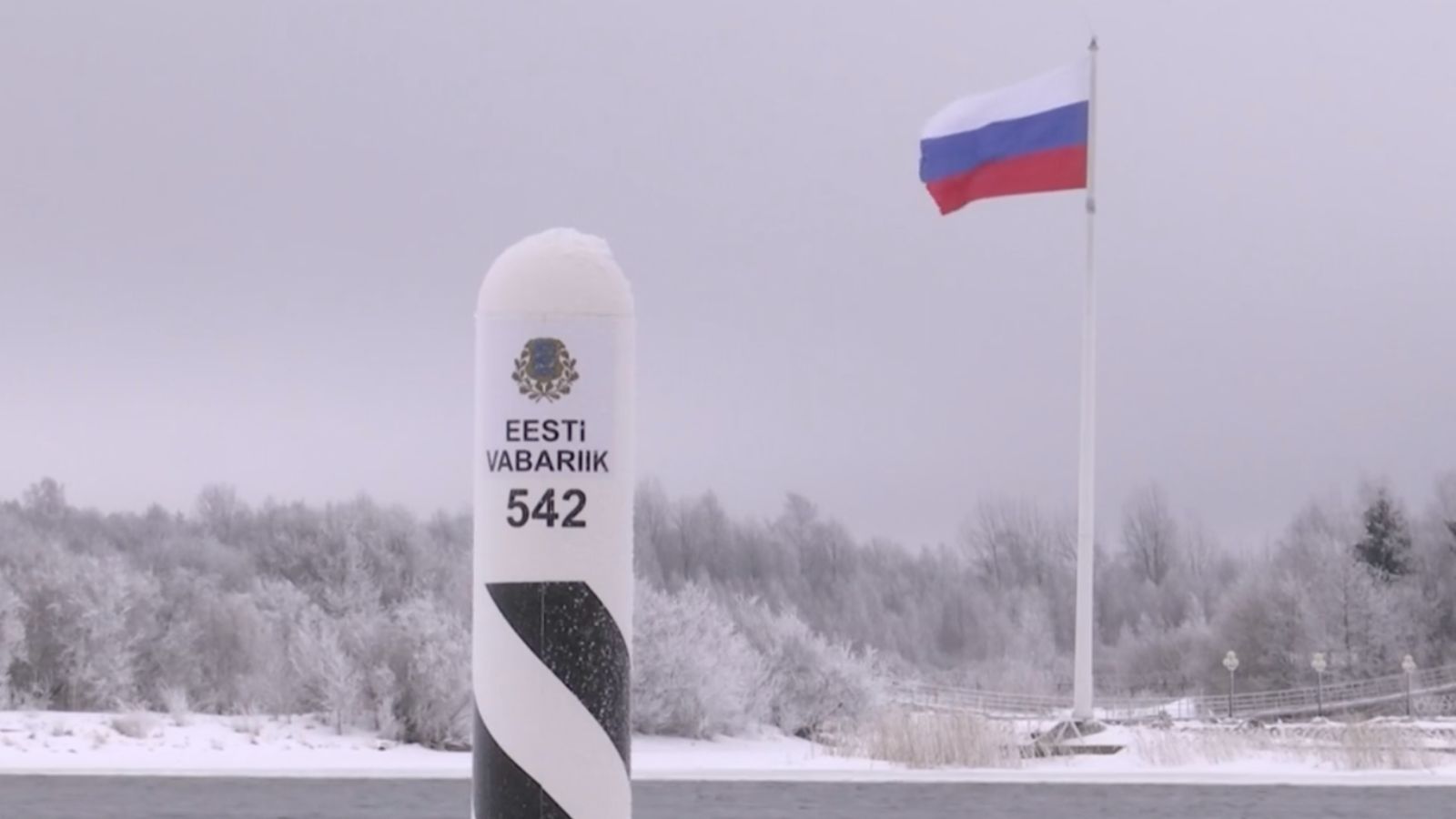 Estonian border post with the national coat of arms against a winter landscape and a Russian flag in the distance.