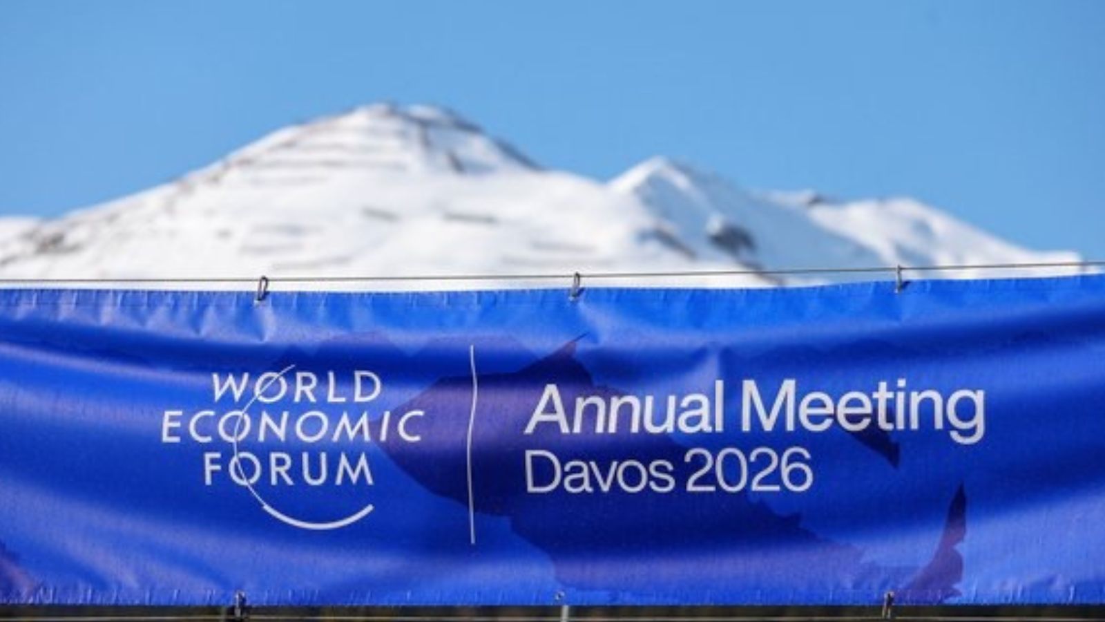 A blue horizontal banner for the World Economic Forum Annual Meeting Davos 2026 stretched across the foreground, with a snow-covered mountain peak visible under a clear blue sky in the background.