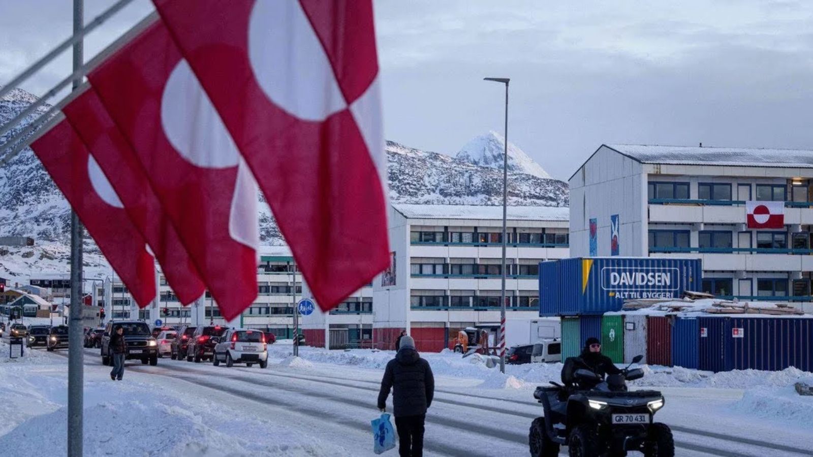 A snowy street in Greenland featuring Greenlandic flags in the foreground and residential buildings against a mountain backdrop.