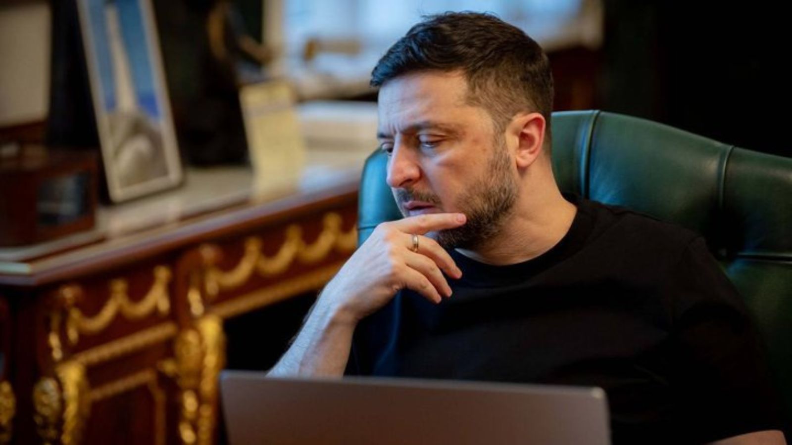 A portrait of Volodymyr Zelenskyy looking intently at a laptop screen with his chin resting on his hand
