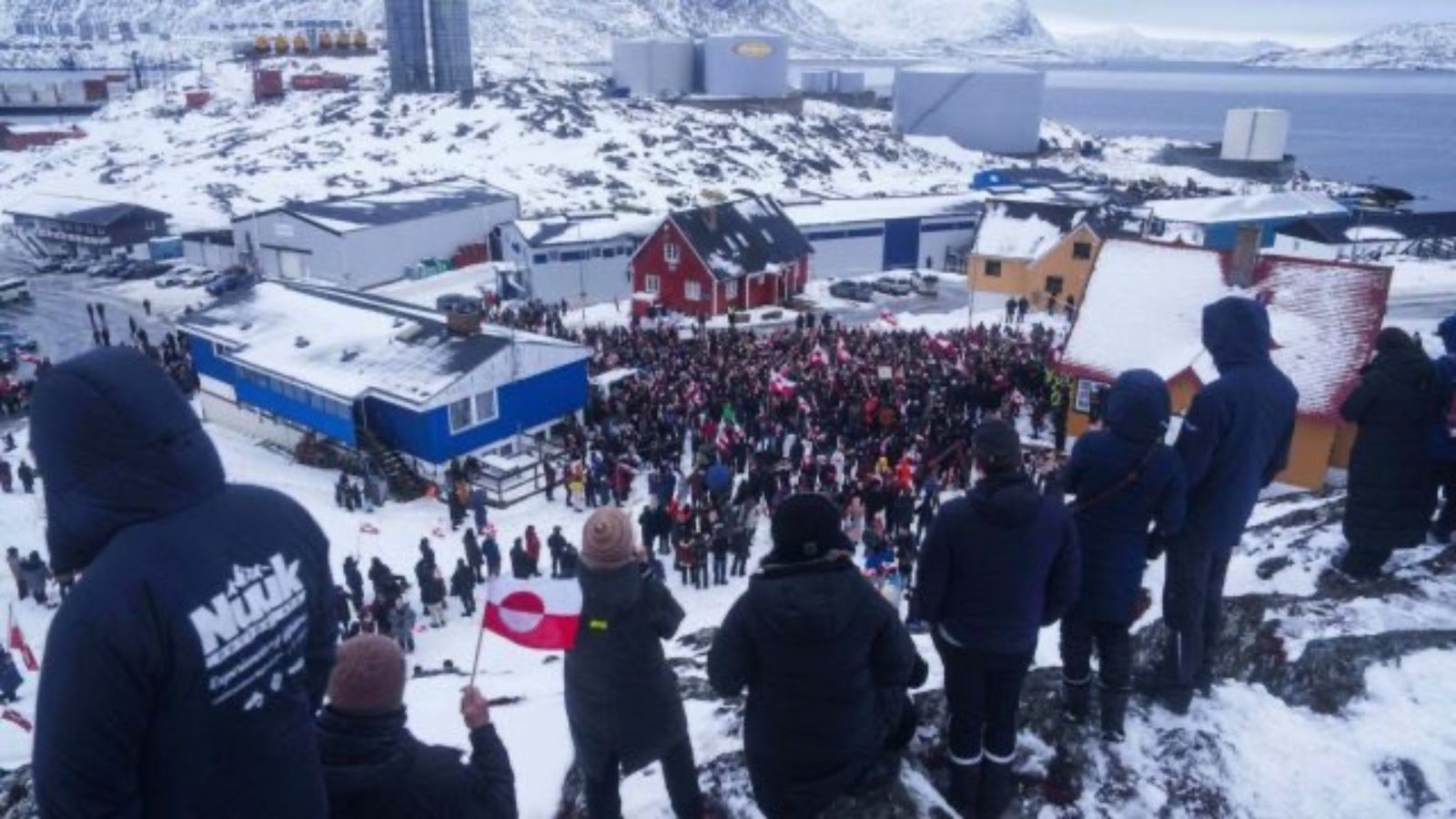 A large crowd of people gathered in a snowy town square in Nuuk, Greenland, holding flags and protesting, viewed from a high rocky vantage point with residential buildings in the background.