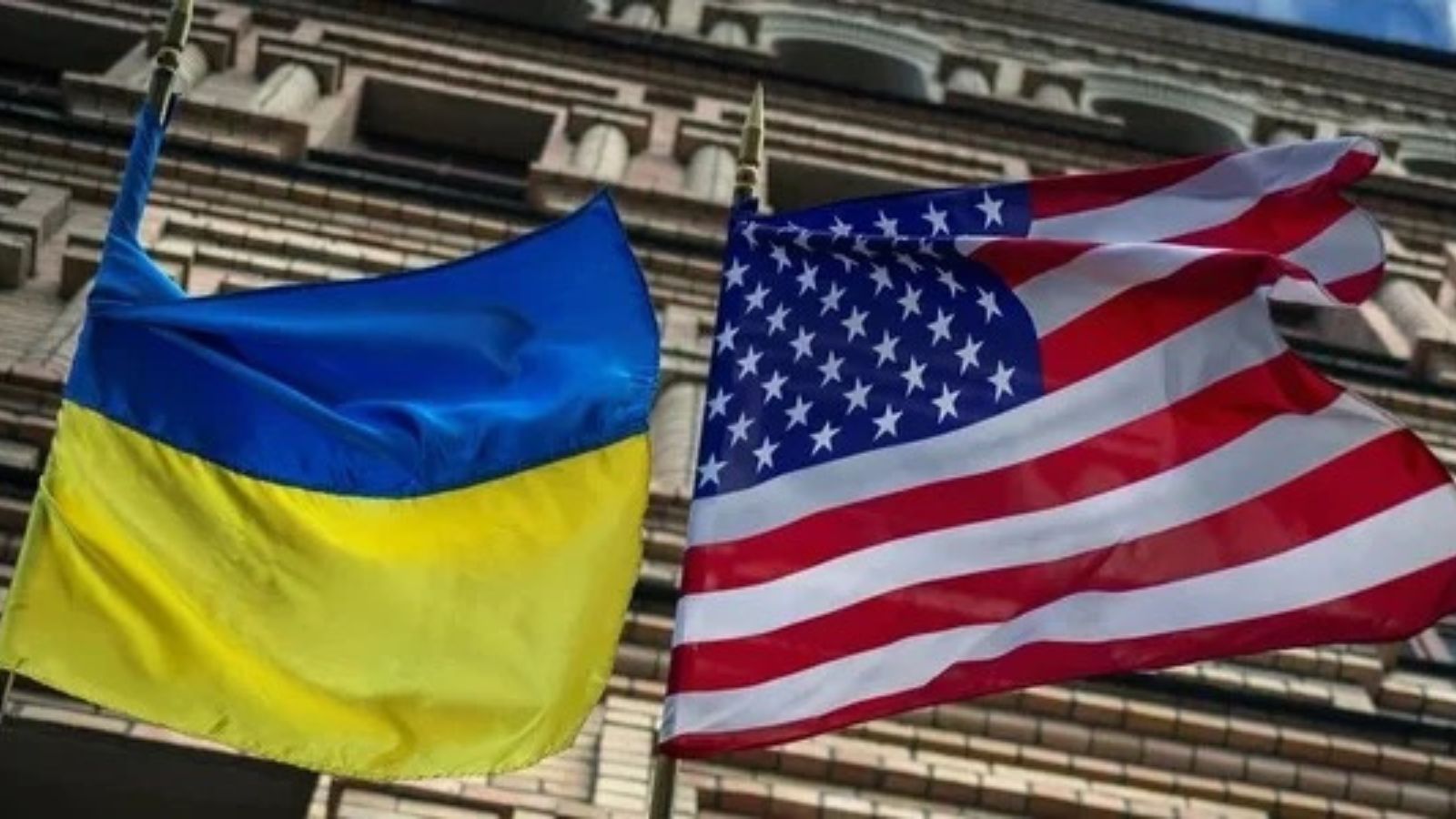 Ukrainian and United States flags waving side by side in front of a brick building.