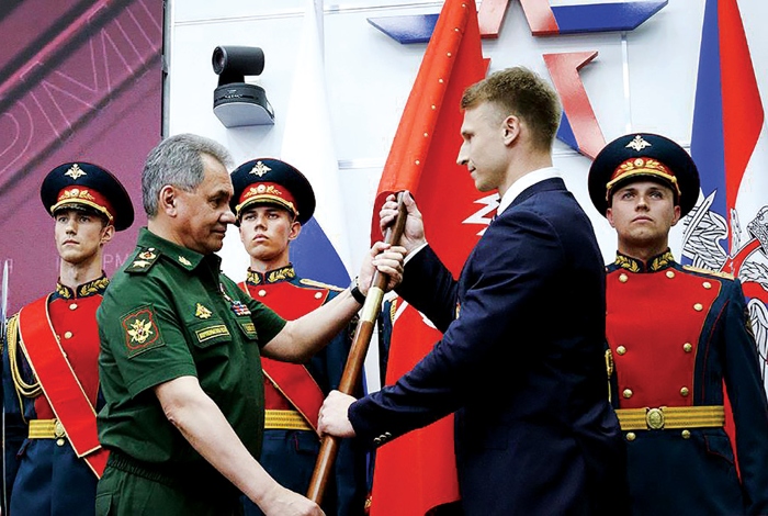 A Russian high-ranking official in military uniform hands over a flag to a young member of a military-patriotic organisation during a solemn ceremony.