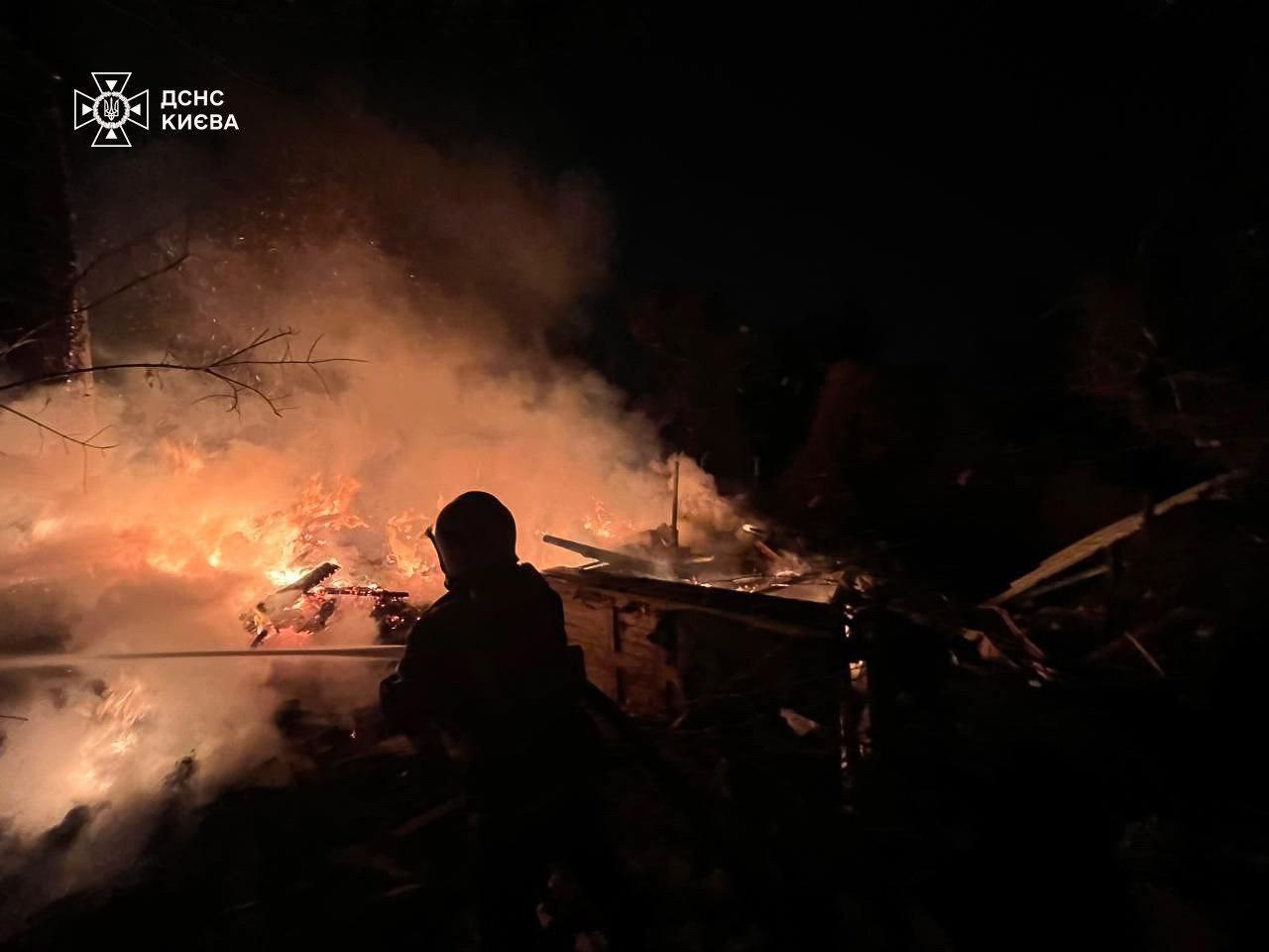 Ukrainian emergency responder battles a large nighttime fire amid debris and smoke after a russian attack on Kyiv, with flames illuminating the scene as firefighters work to extinguish burning structures following shelling or drone strikes.