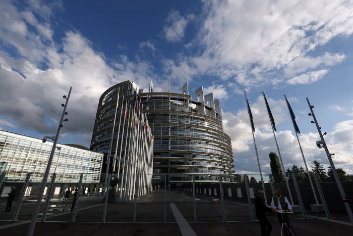 Exterior view of the European Parliament building in Strasbourg under a partly cloudy sky, with EU flags flying in front of the modern glass-and-steel structure.